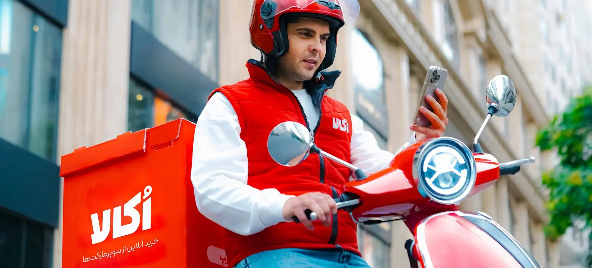 سفیر اکالا در حال بررسی مسیر تحویل سفارش سوپرمارکتی با موتور قرمز – ارسال رایگان، خرید به‌صرفه، پشتیبانی عالی | Okala delivery rider checking route on red scooter – Free delivery, affordable shopping, excellent support