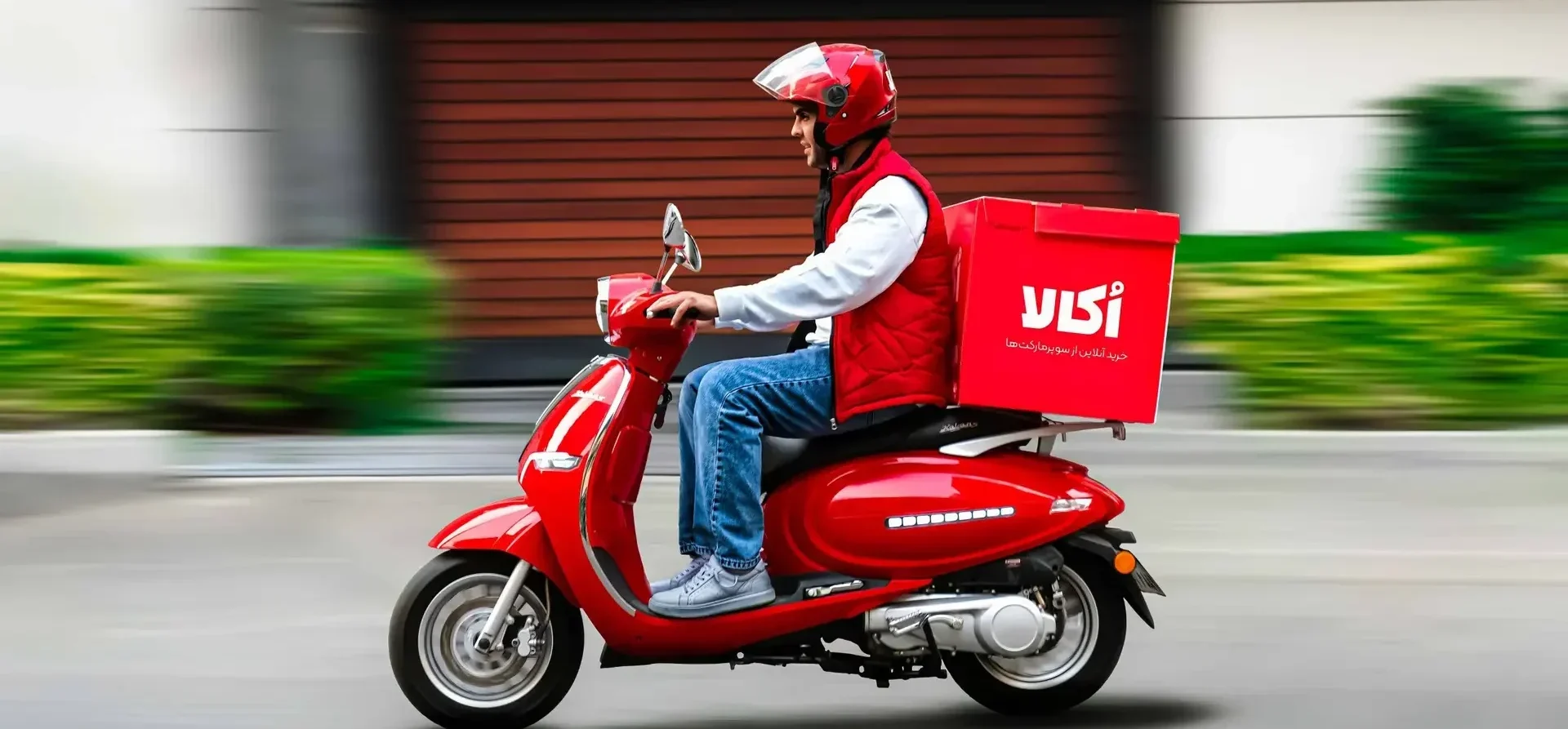 سفیر اکالا در حال تحویل سفارش سوپرمارکتی با موتور قرمز – ارسال رایگان، خرید به‌صرفه و پشتیبانی عالی | Okala delivery rider with red scooter – Free delivery, affordable shopping, excellent customer support